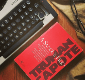 livro de capa vermelha com o nome do autor em preto. ao lado, uma máquina de escrever cinza. aparece ainda um par de óculos e um tampo de madeira por baixo de tudo