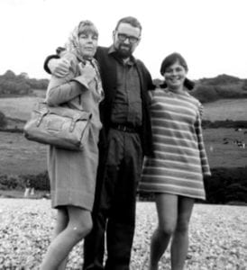 Elizabeth Fowles, John Fowles e a filha de Elizabeth Anna Christy, nos arredores de Underhill Farm, Lyme Regis, Dorset, verão de 1966. eles estão abraçados, a essa esta de vestido e segura uma bolsa e esta com um lenço na cabeça, Fowles esta de casaco es a filha esta com um vestido listrado. eles estão no campo.