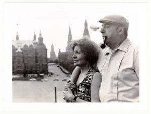 foto em preto e branco de Neruda com Matilde Urrutia, Eles de perfil, com prédios com pináculos ao fundo. Ele de boina e fumando charuto