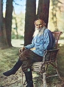 Tolstói, bem idoso, com barba longa e branca, sentado num banco de parque, vestindo calça marrom, camis azul e botas pretas