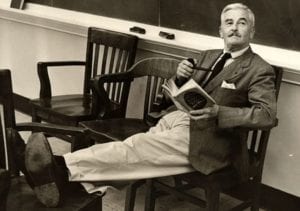 Autor sentado em uma cadeira com as pernas esticadas e apoiadas em outra cadeira com um livro nas mãos 